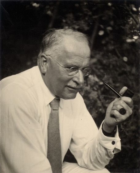 Carl Jung sitting outdoors with a pipe, wearing glasses and a tie, in a reflective pose.