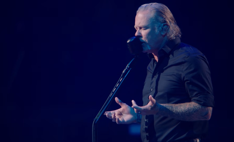 Close-up profile of James Hetfield singing passionately into a vintage microphone with open, outstretched hands in deep blue stage lighting.