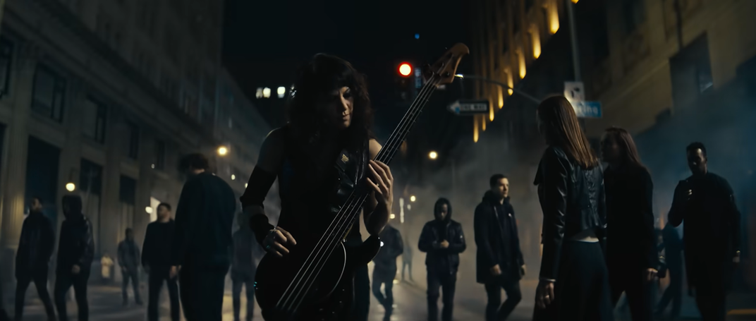 Bassist, Emma Anzai, playing in the middle of a dark city street surrounded by figures in the Who Will You Follow video.