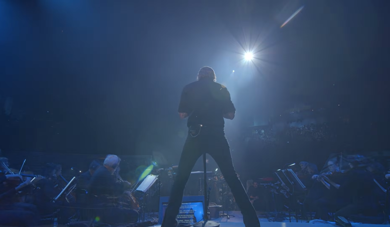 Back view of James Hetfield standing at a microphone on stage, backlit by a bright overhead spotlight while surrounded by the San Francisco 