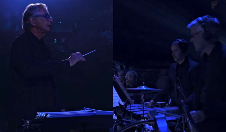 Split-view shot of an orchestra conductor holding a baton in blue light alongside percussionists playing behind a music stand.